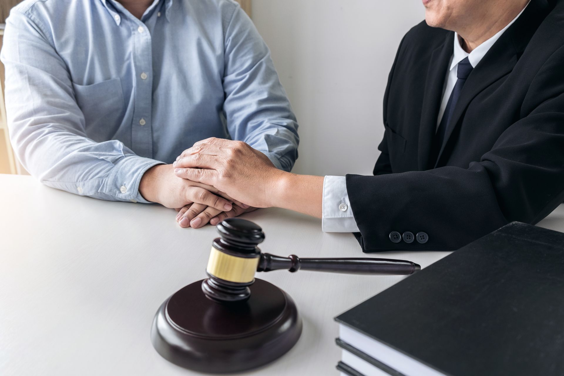 Lawyer consoles client at a table with a gavel and law book; hands clasped in support.