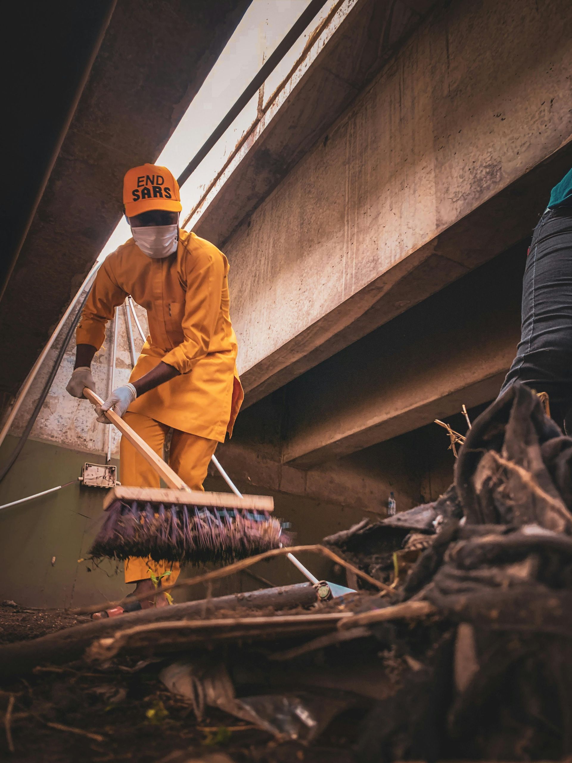 Person in orange uniform sweeps debris under a bridge; mask, hat, and gloves.