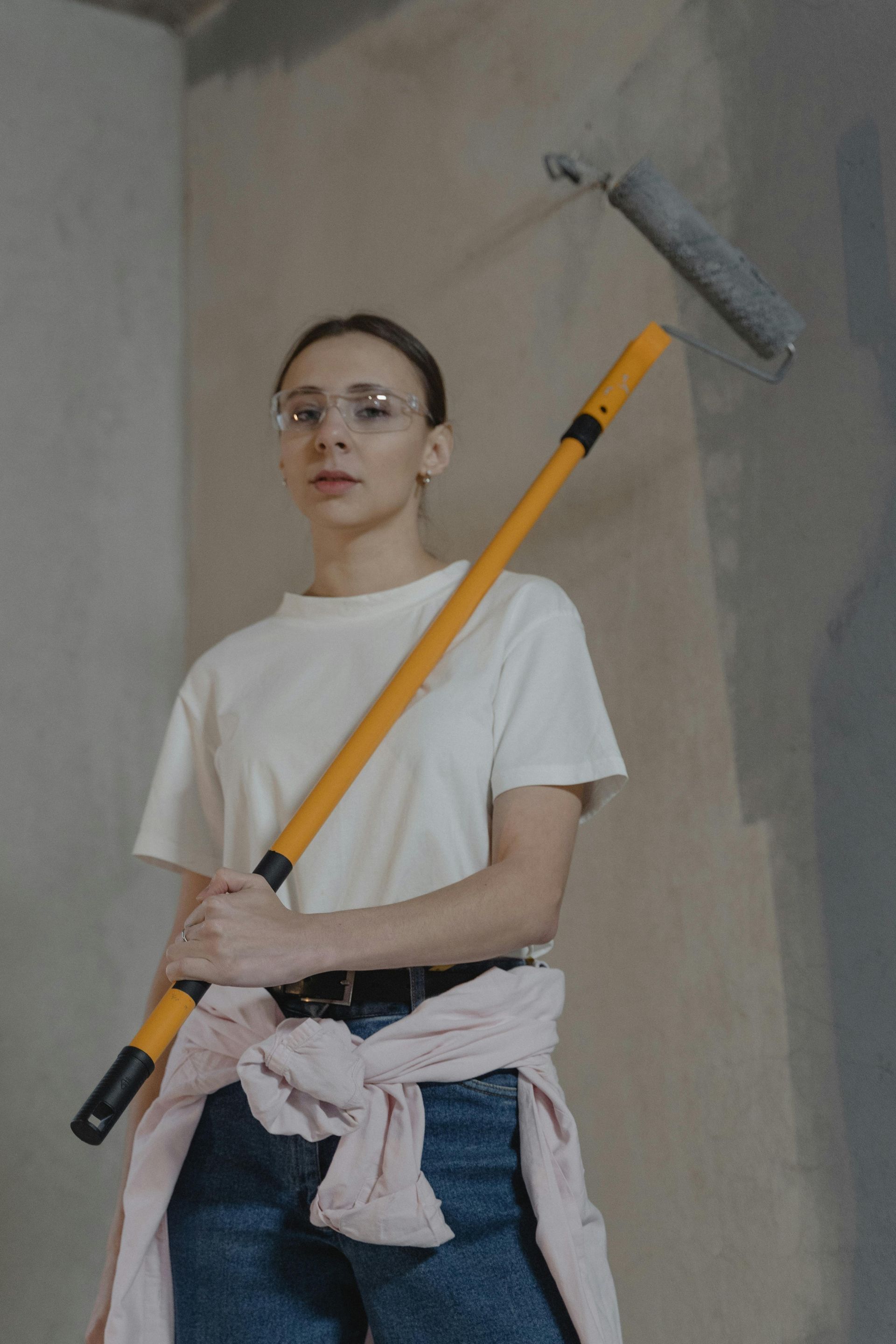 Woman in glasses with paint roller; interior construction, neutral color.