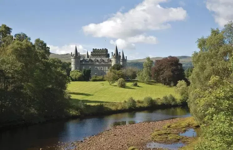 A castle in Scotland with spired turrets sits on a grassy hill beside a river under a blue sky with clouds.