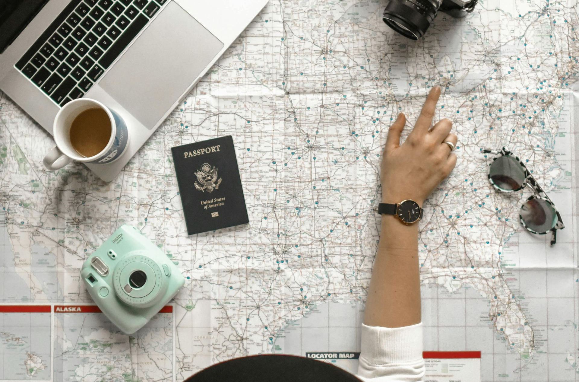 Person's hand pointing at map with travel items: laptop, camera, passport, sunglasses, coffee.