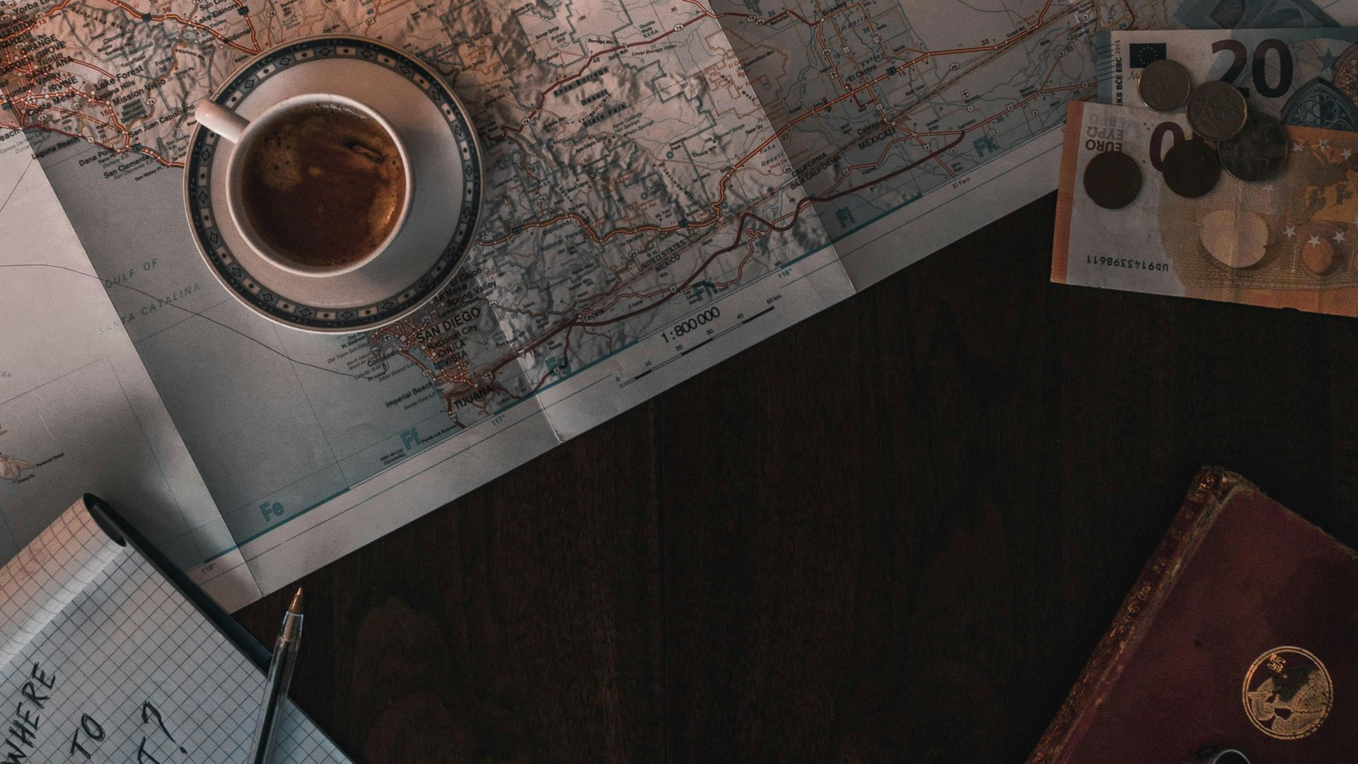 Coffee cup on a map with money, notebook, and journal on a wooden surface, suggesting travel planning.