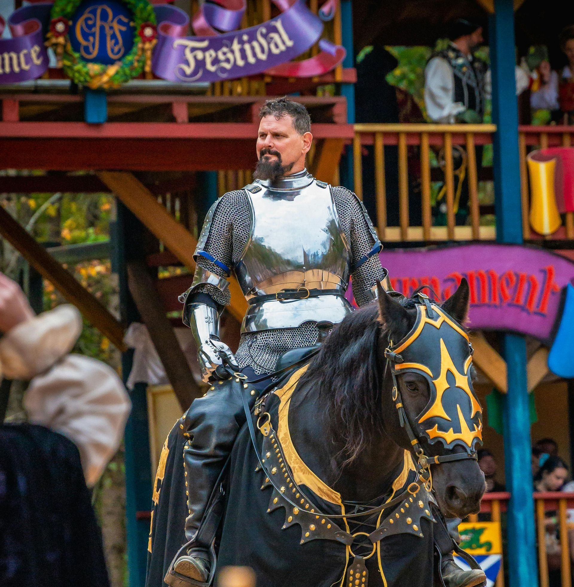 A man in silver armor rides a black horse at a Renaissance festival. He has a mustache, is smiling, and the horse has gold and black decorations.