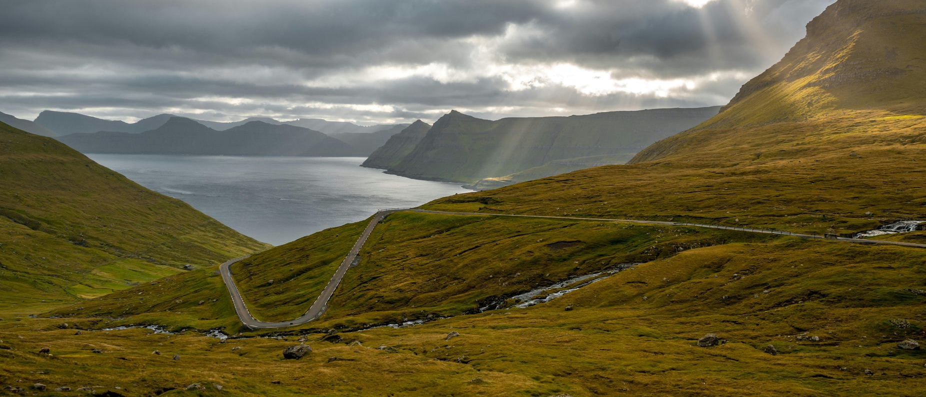 A winding road leads through a valley to a body of water under a cloudy sky, in Norway