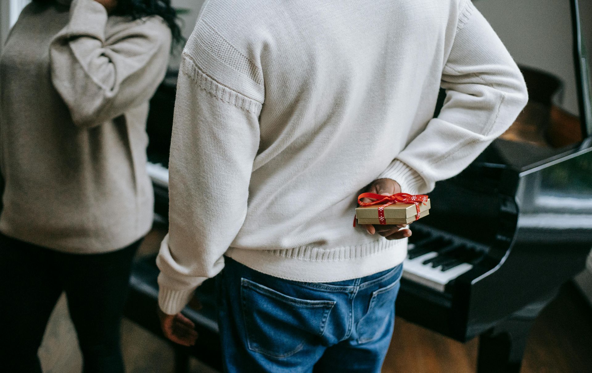 Person holding wrapped gift behind back, near piano, with another person in the background.