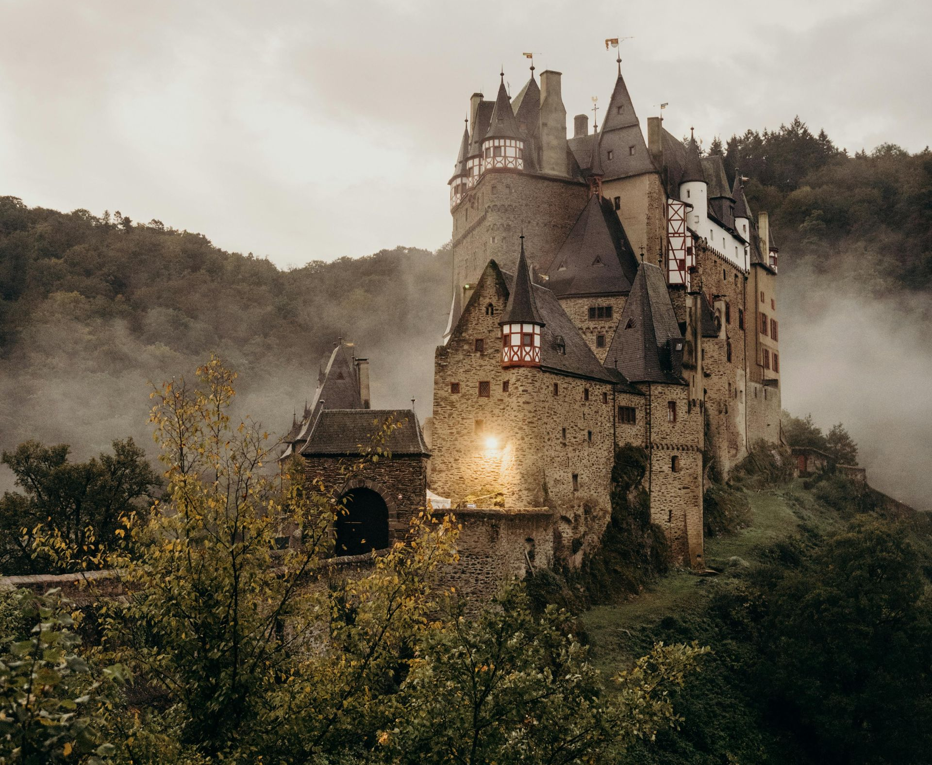 Stone castle on a hilltop shrouded in mist with a cloudy sky and surrounding trees.