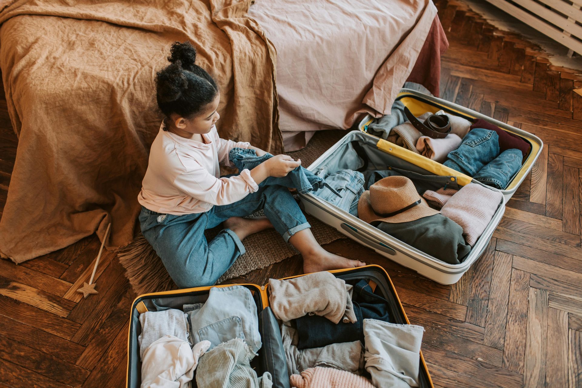 Person packing clothes in open suitcases on a wooden floor near a bed.