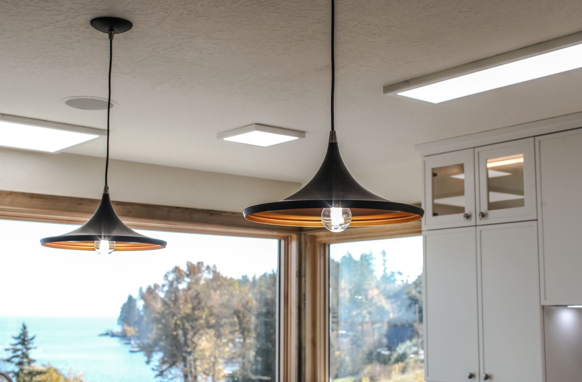 Two black pendant lights hanging above a kitchen counter with a window and white cabinets.