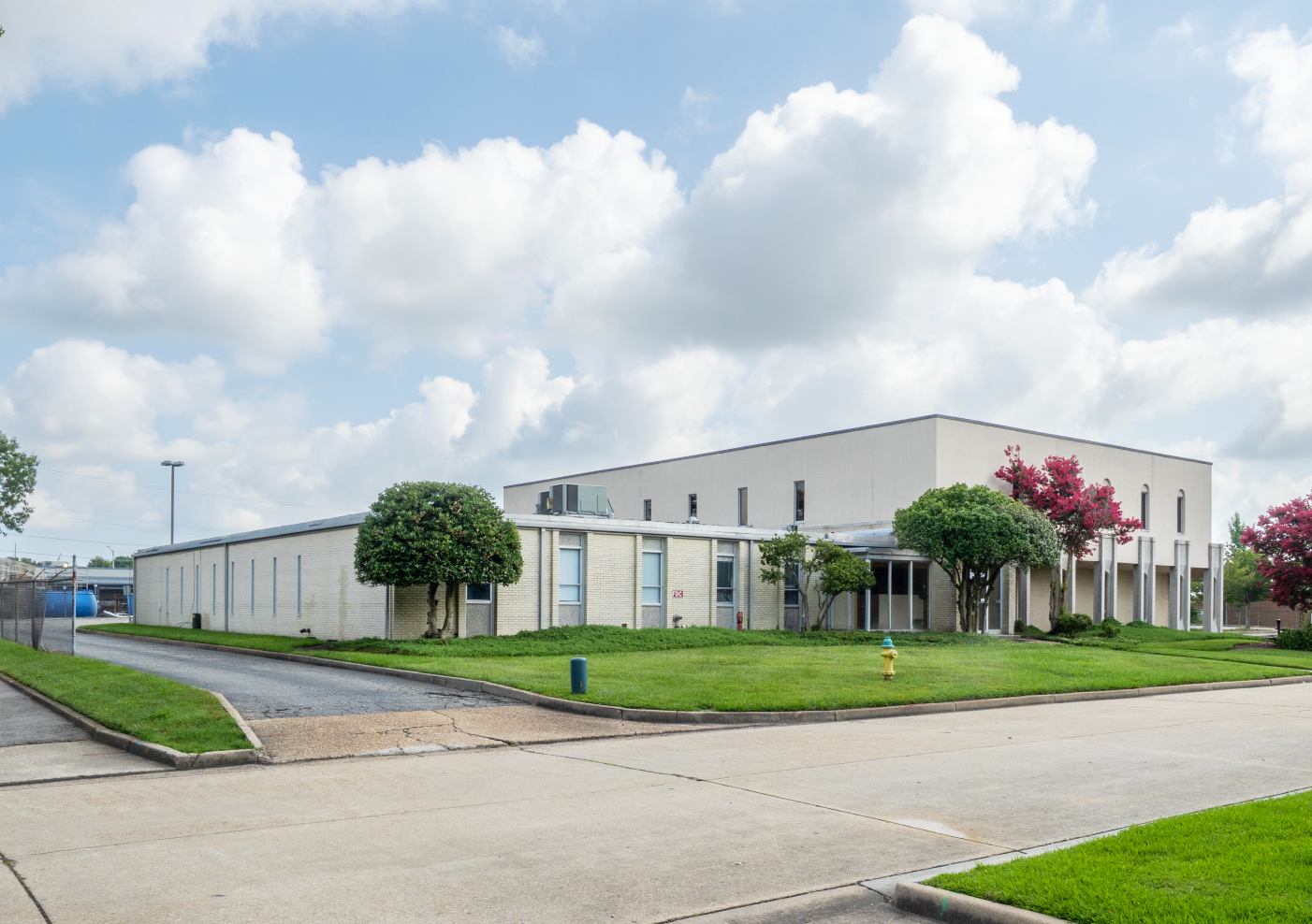 Low-rise, light-colored industrial building with trees in front on a sunny day with a blue sky and clouds.