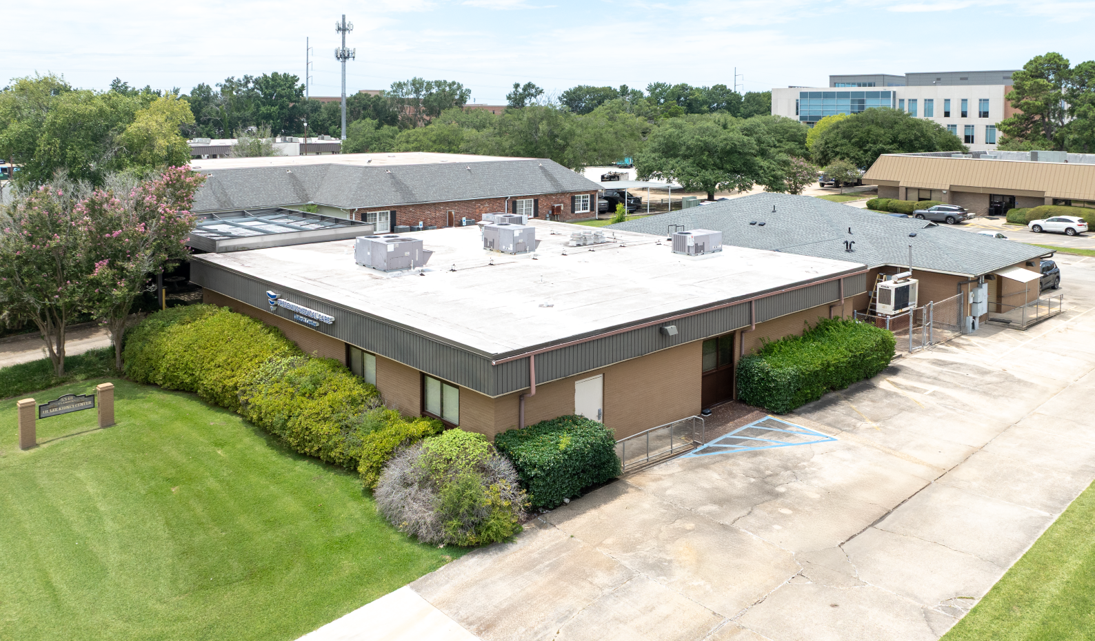 An aerial view of a one-story brick building with a flat roof, surrounded by green bushes and grass, in a commercial area.