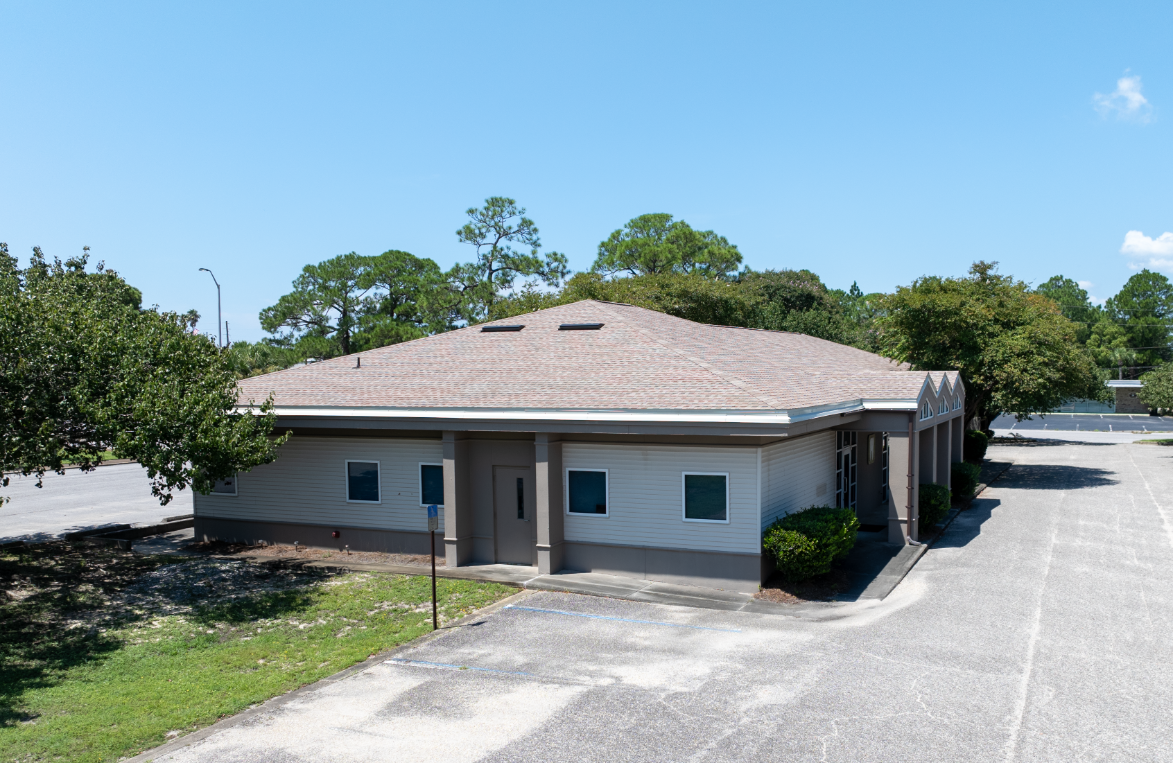 Single-story tan building with brown tile roof; small windows and dark door; surrounded by trees and parking lot; clear blue sky.
