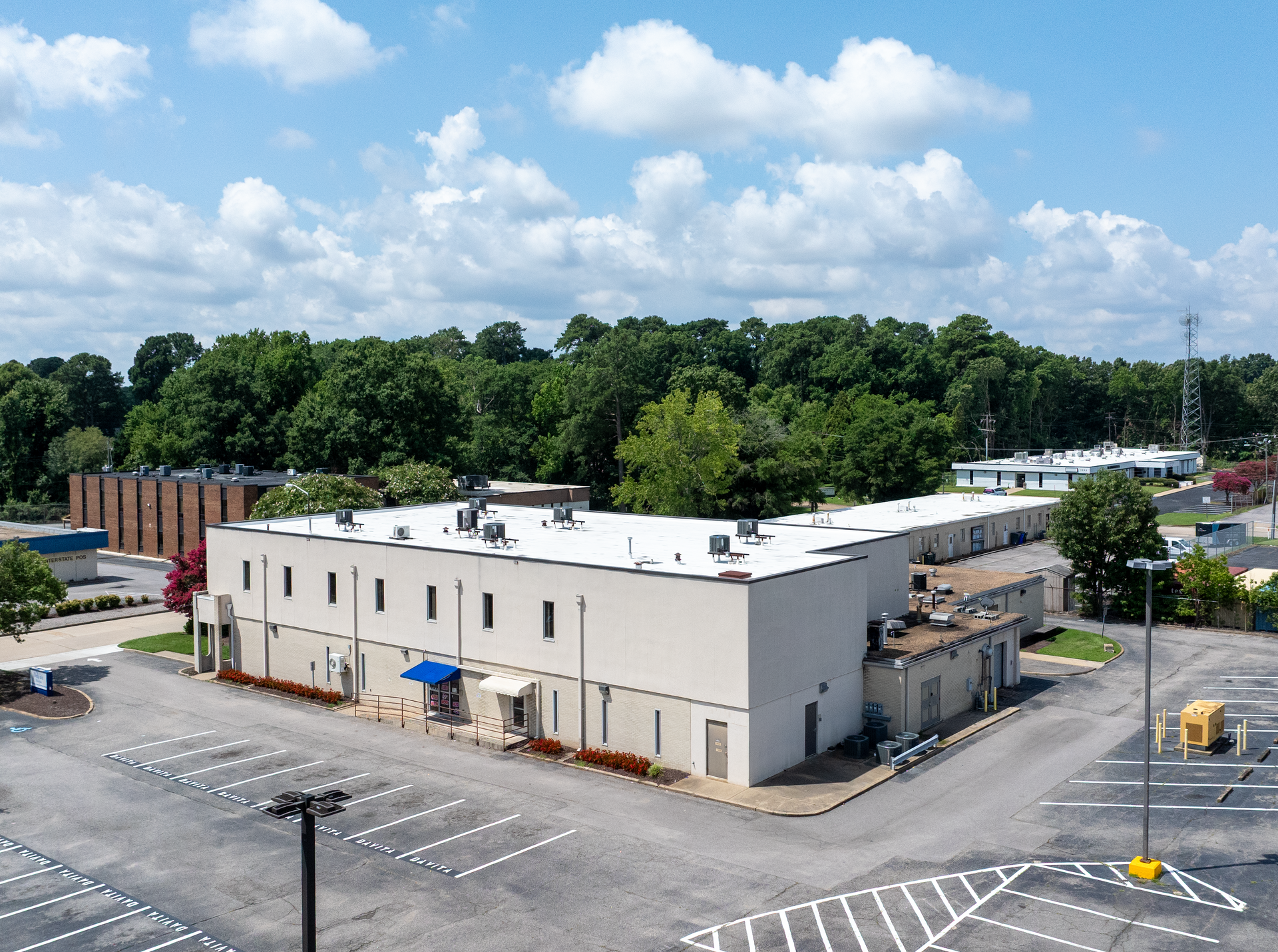 An aerial view of a two-story beige building with a flat roof and surrounding parking lot, green trees, and a blue sky.