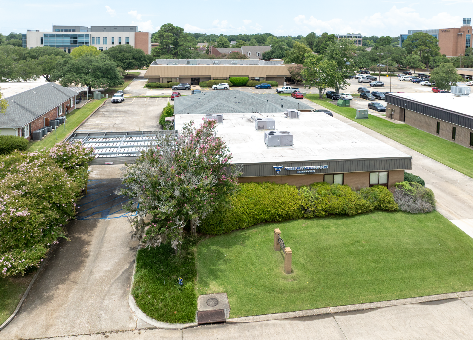 Exterior view of a single-story building with a flat roof, green lawn, and small trees. Cars parked in the background.
