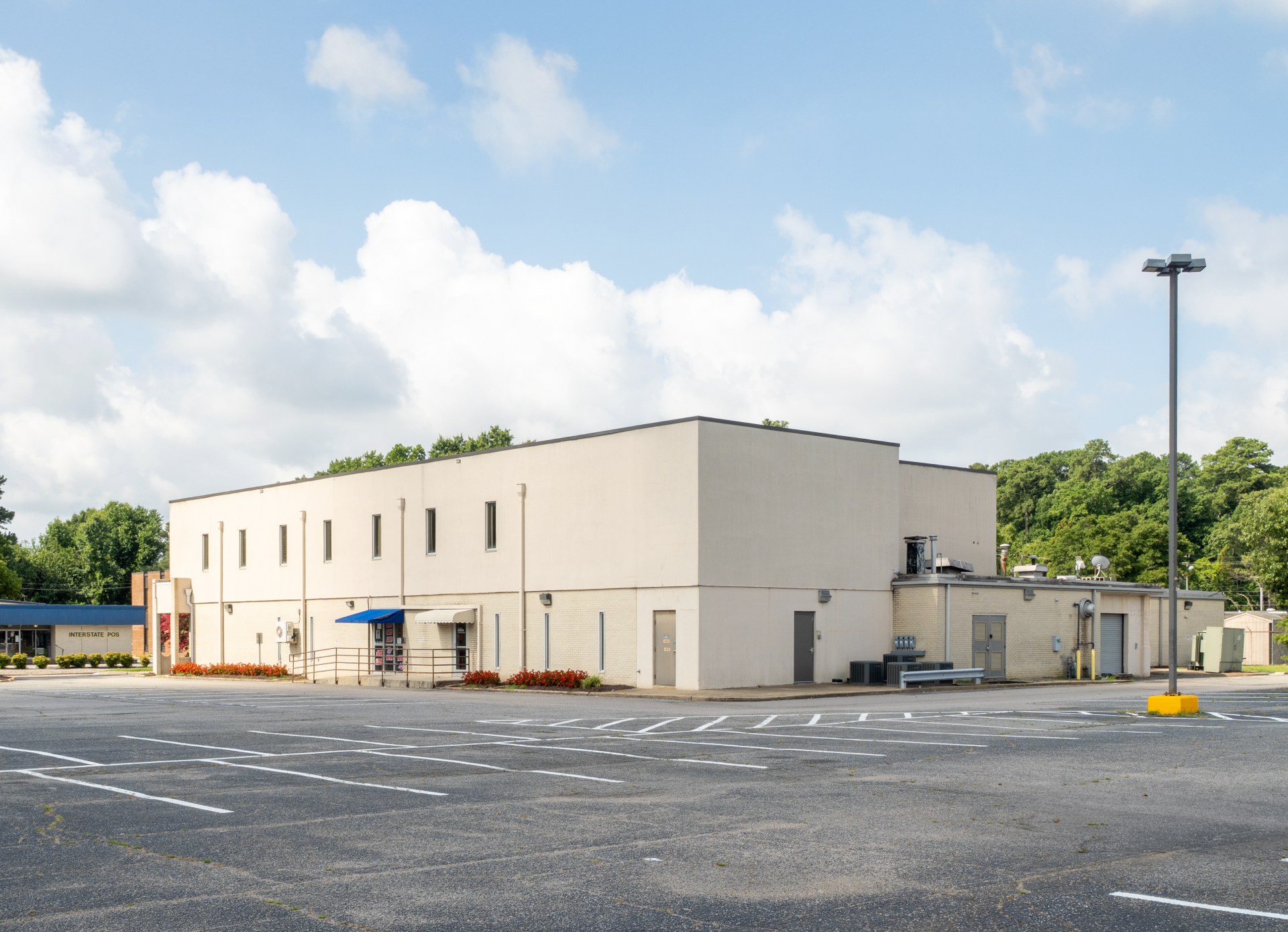 A large, off-white commercial building with a mostly empty parking lot. Sunny day with some trees in the background.