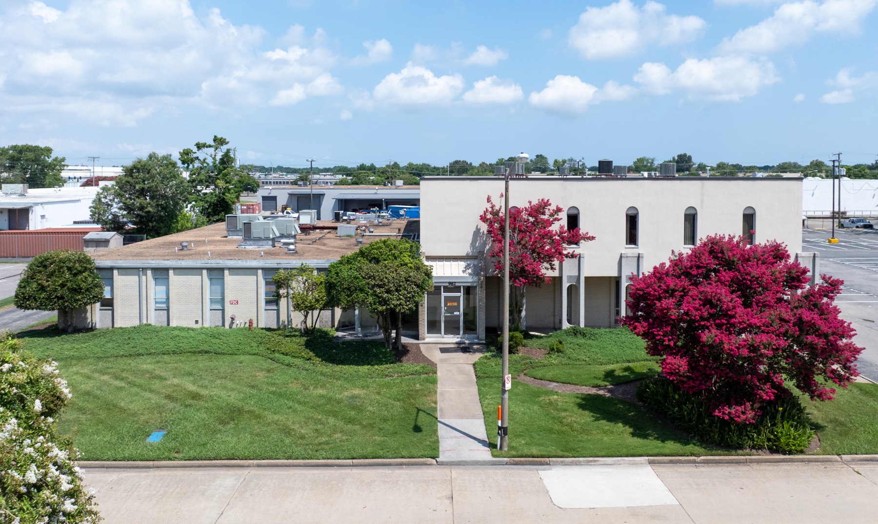 Exterior view of a light-colored, two-story commercial building on a sunny day, with a paved path leading to the front entrance. Lush, red-leafed tree on the right side of the building.