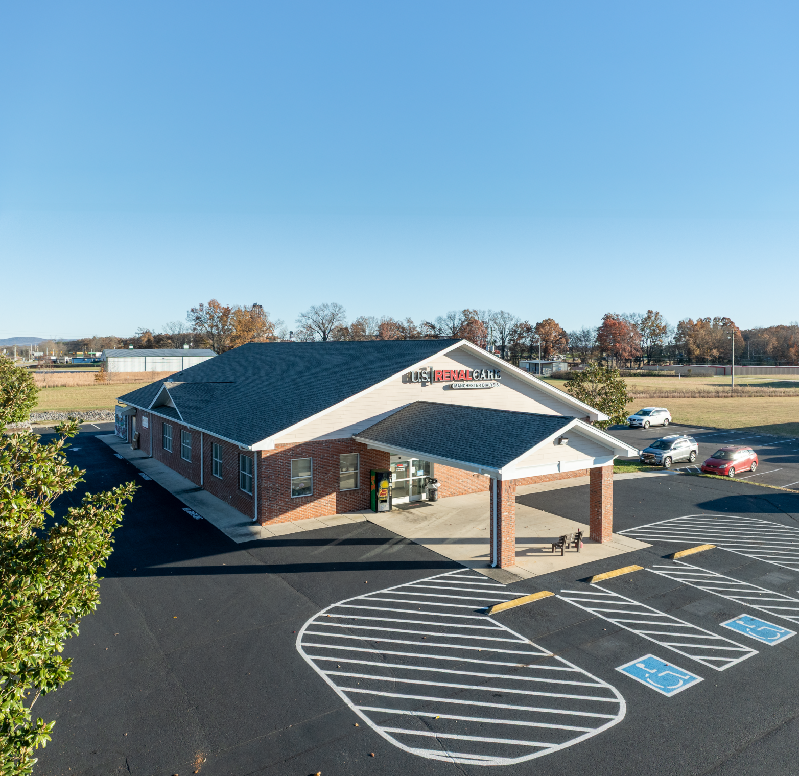 US Renal Care net lease medical dialysis building with a dark roof and covered entrance. Cars parked in front. Blue sky.