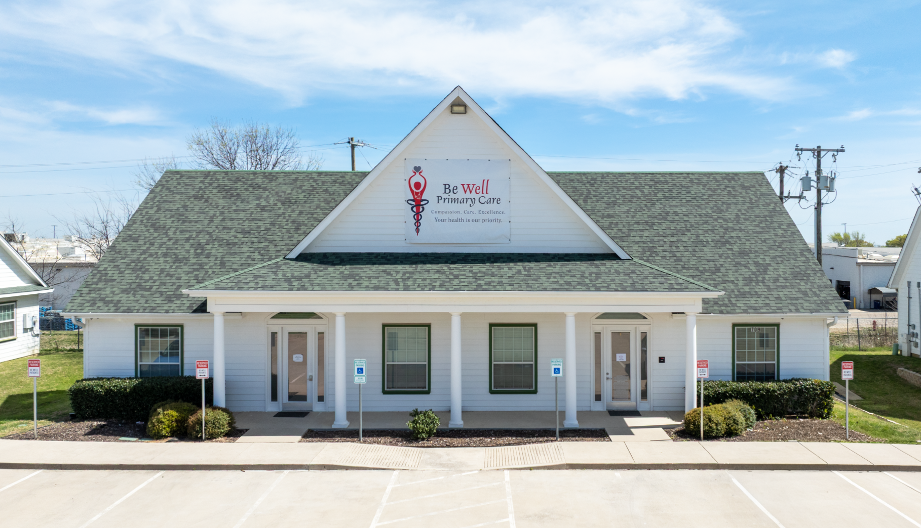 A front-facing view of a white, single-story BeWell Primary Care medical building with a gray roof and a parking lot in front.