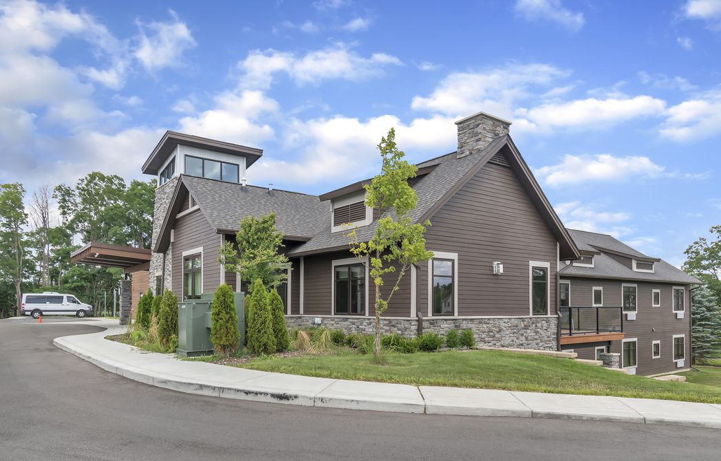 A brown building with a stone facade, tower, and chimney sits on a slight hill under a blue sky. A curved driveway leads to the entrance.
