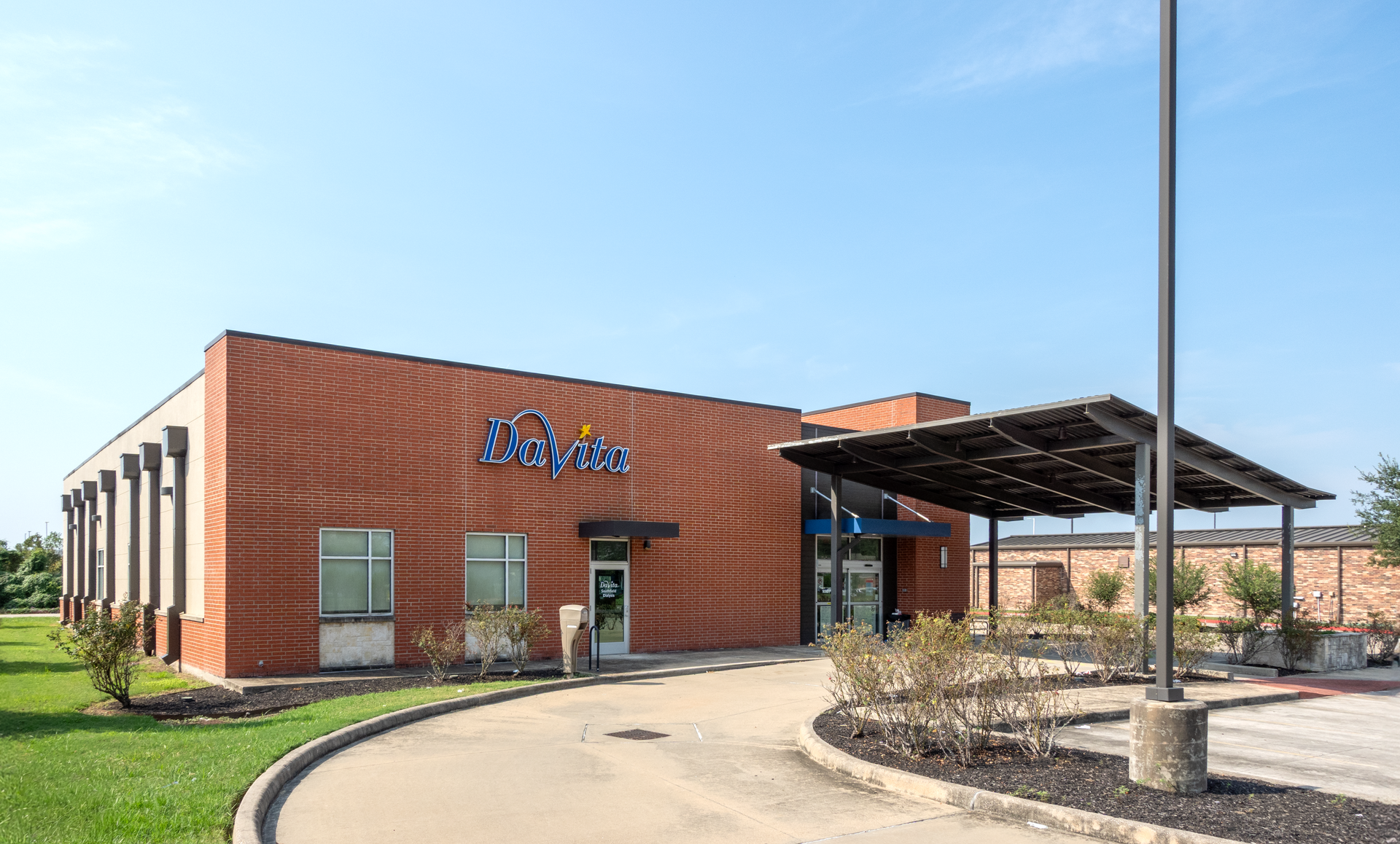 Red brick Davita dialysis building with blue sign, canopy, and pathway leading to the entrance under a clear, blue sky.