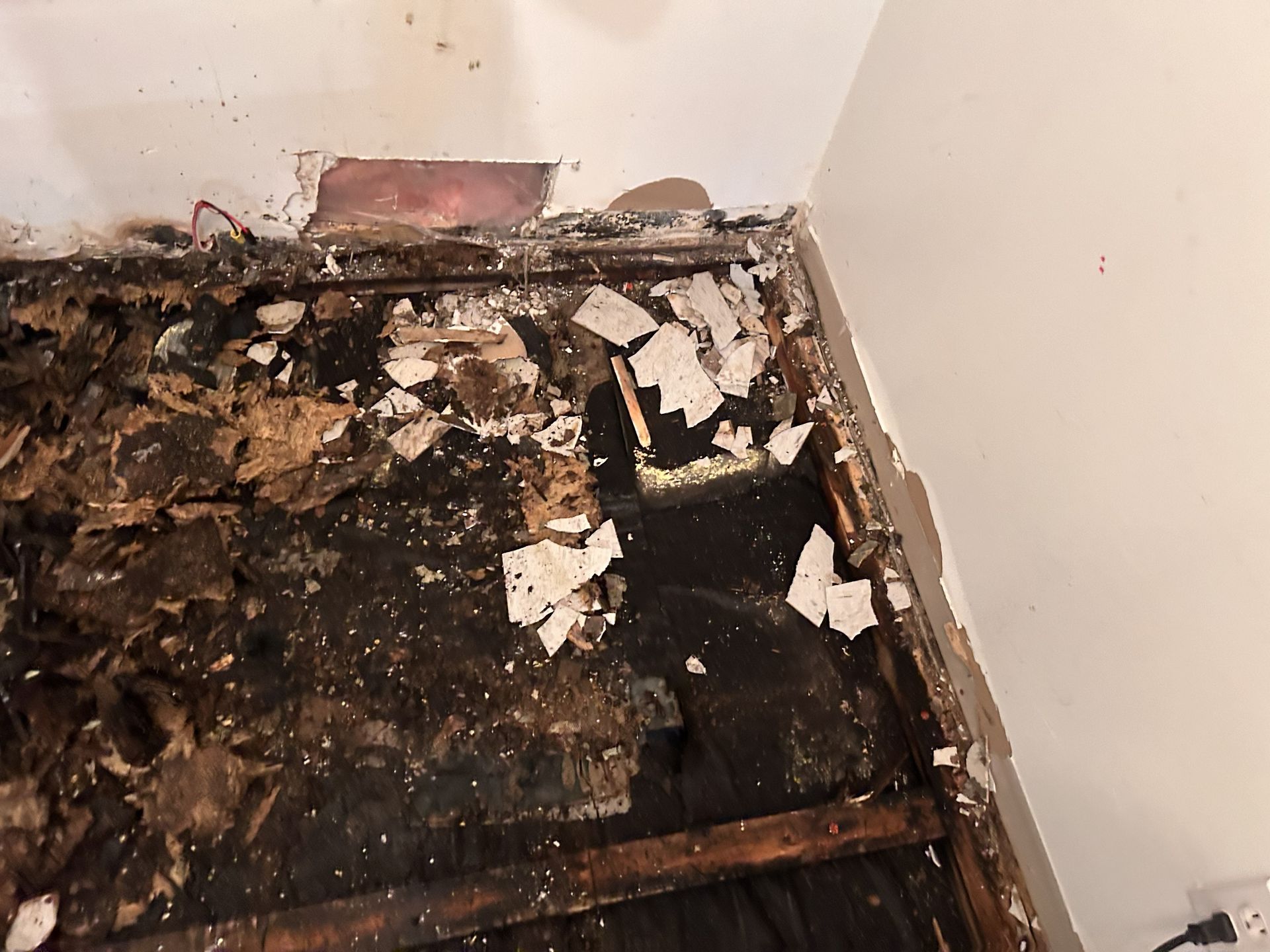 Un coin de pièce avec un sol noir arraché et endommagé par l'eau, et des débris à côté d'un mur avec des plaques de plâtre et du bois apparents.