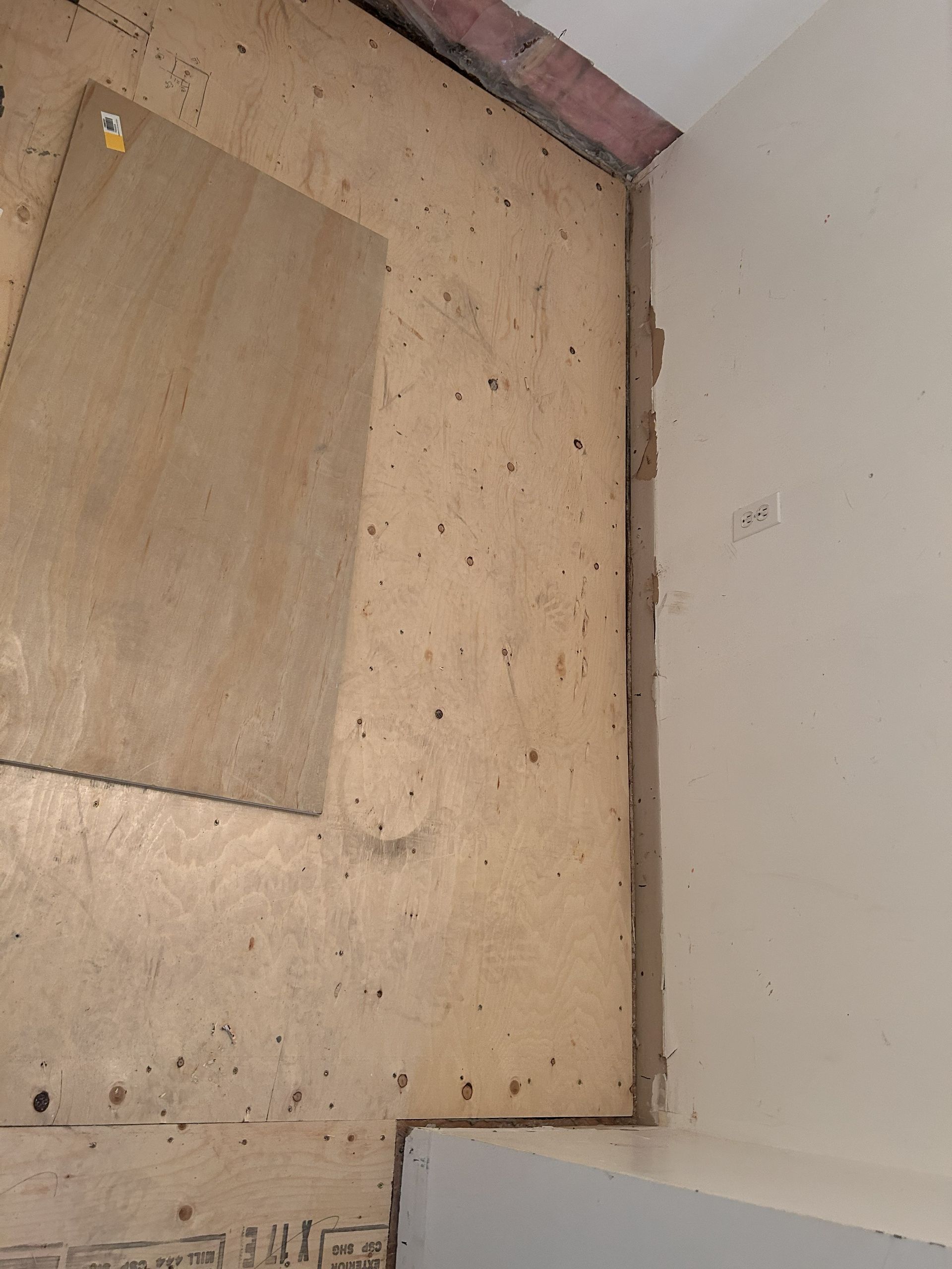 Un sous-plancher en bois en construction, montrant des panneaux de contreplaqué, des joints et des fixations métalliques à côté d'un mur intérieur simple.