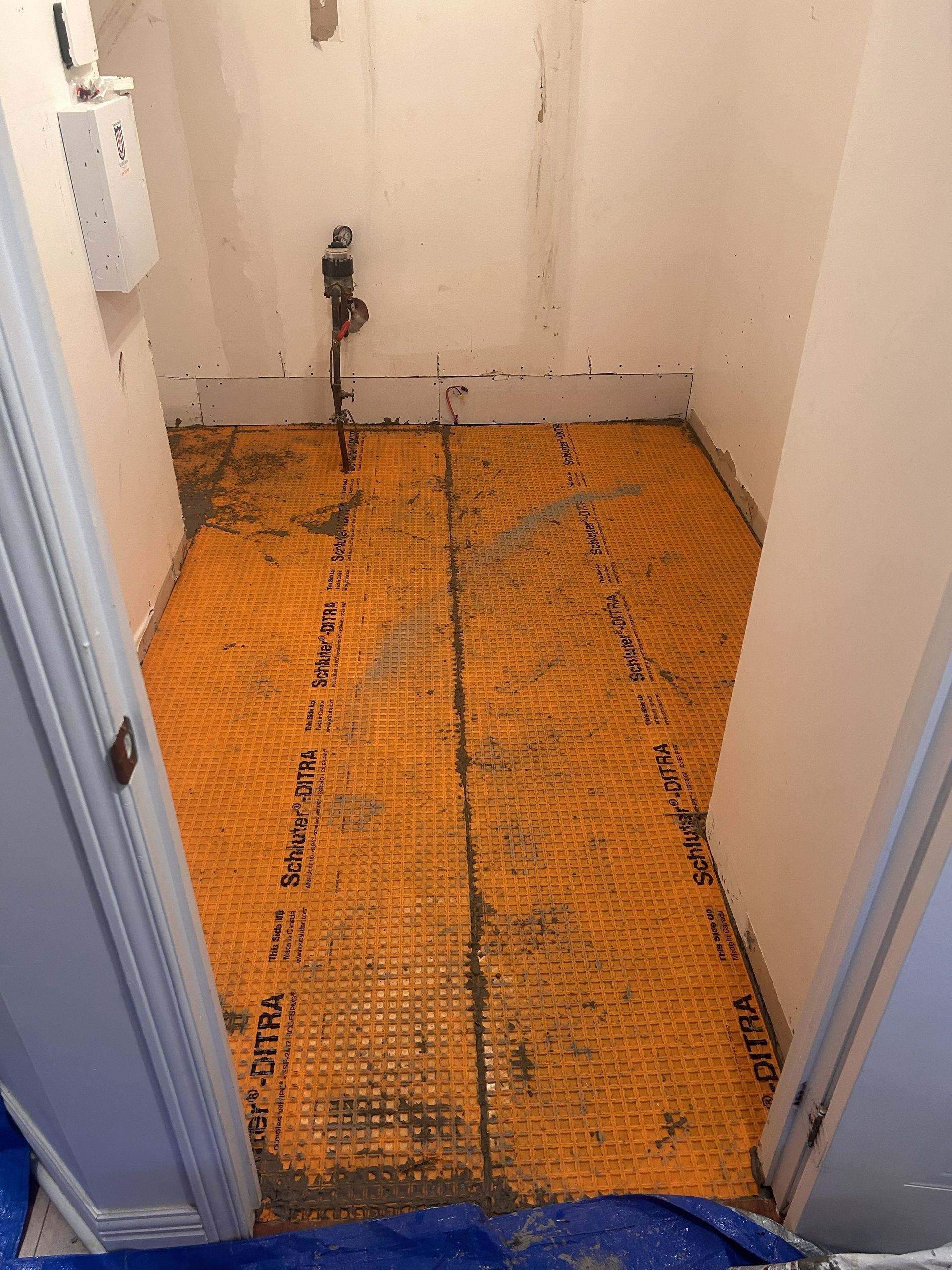 Le sol d'une petite buanderie est recouvert d'une membrane de désolidarisation orange Ditra, avec un tuyau qui dépasse du centre.