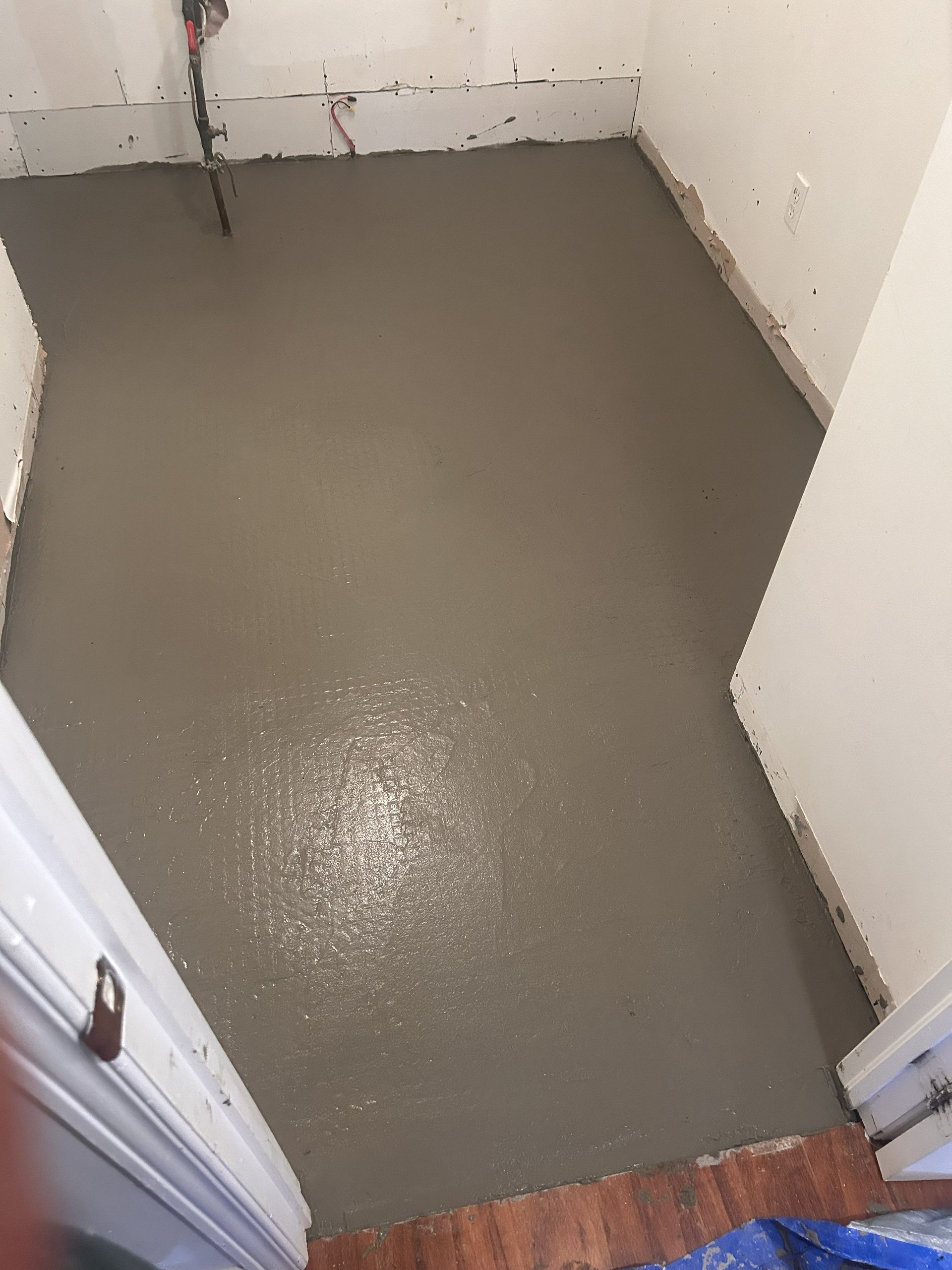 Un sol en béton gris lisse, fraîchement coulé, dans une petite pièce inachevée aux murs blancs et aux fils électriques apparents.