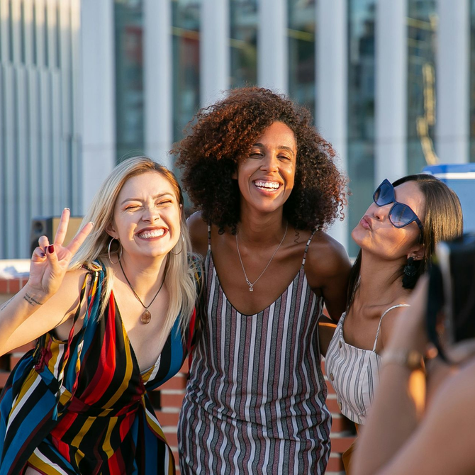 Three smiling women: one makes a peace sign, another puckers lips, and the center woman smiles wide.