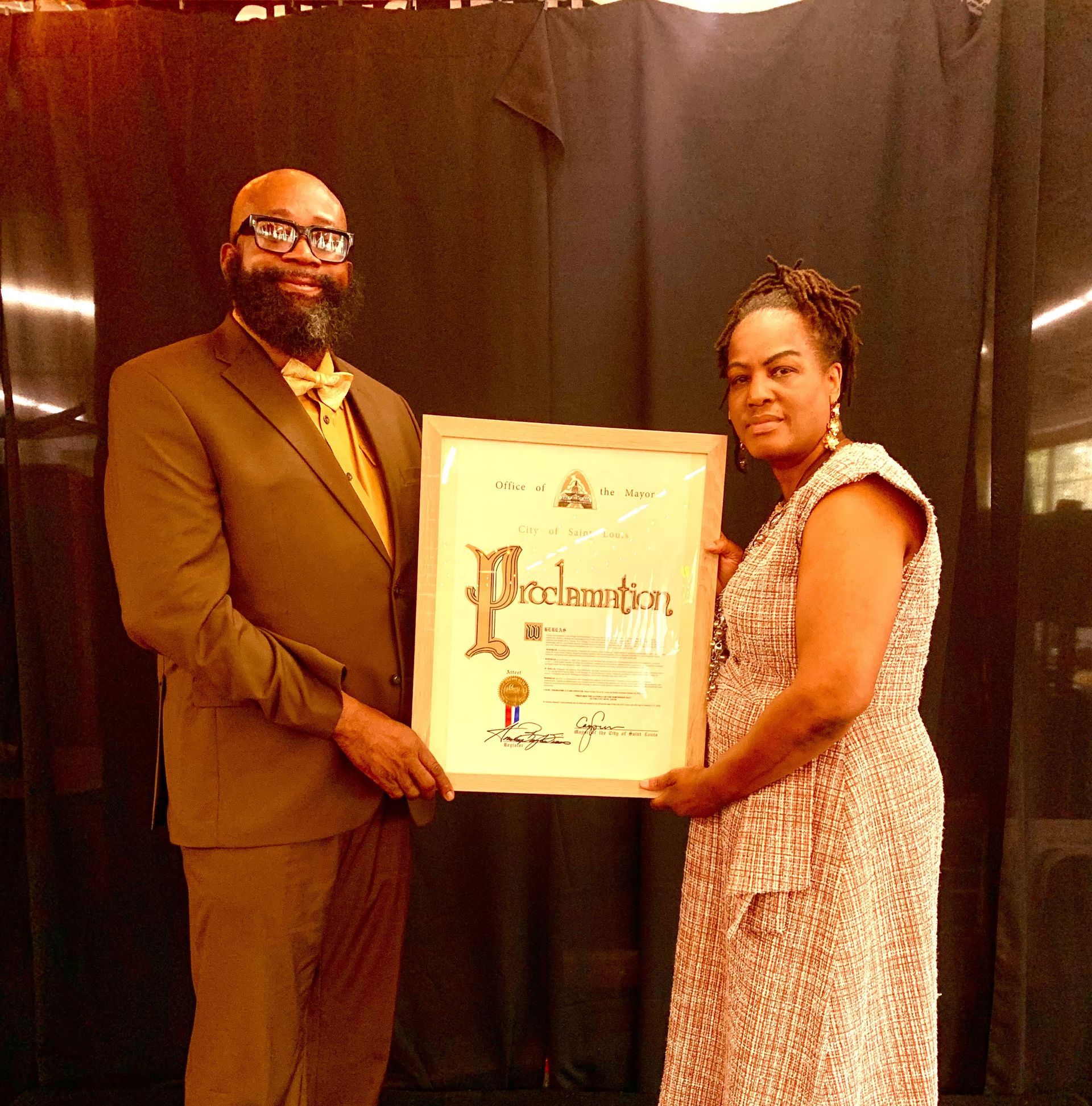 Man in suit and woman holding a framed proclamation. They stand in front of a dark backdrop.