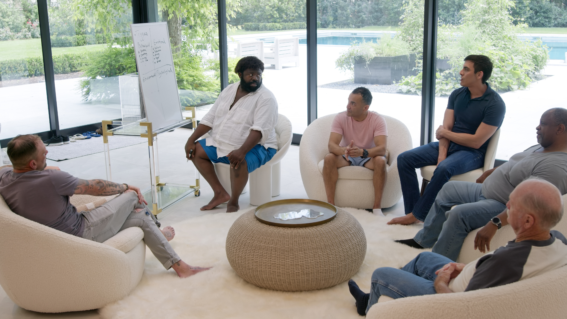 Group of men in a circle, in a sunlit room, engaged in discussion. One man is using a whiteboard.