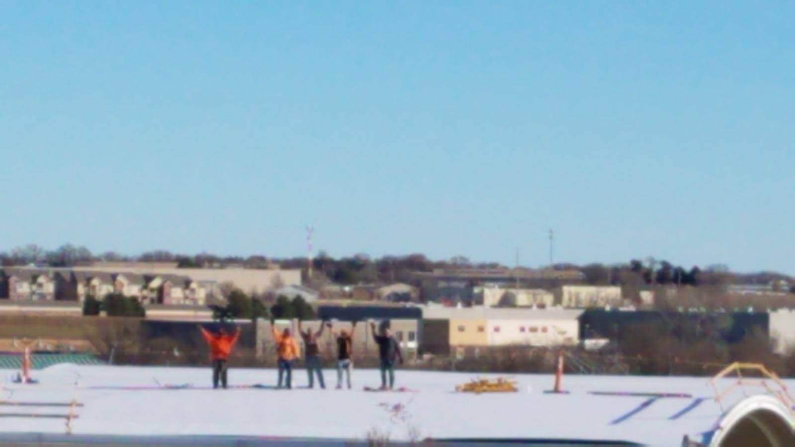 Five people stand on a white rooftop under a clear blue sky, arms raised toward the horizon.