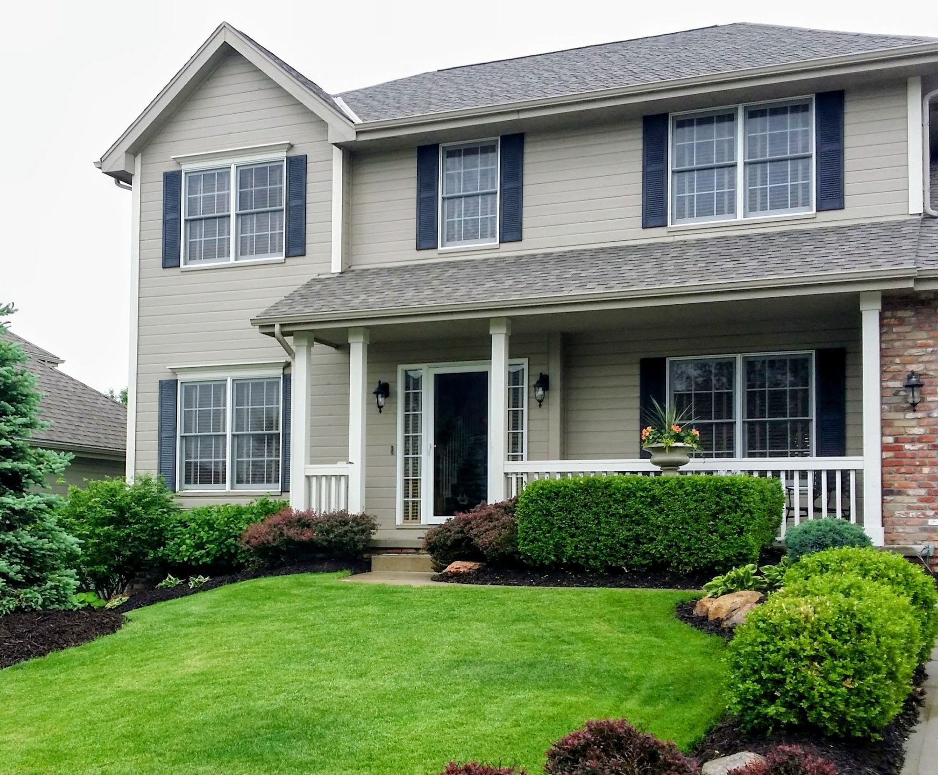 Two-story house with beige siding, dark shutters, a front porch, and a manicured lawn with green bushes.