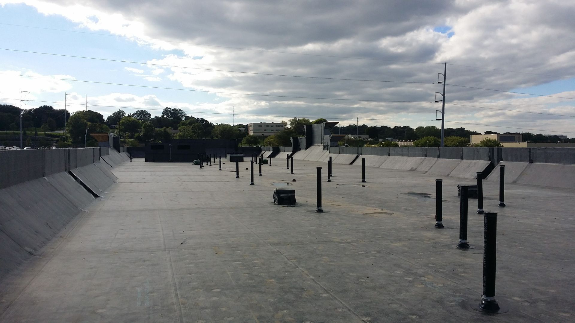 A flat, grey roof under a cloudy sky, featuring numerous vertical black pipes and surrounding low concrete walls.