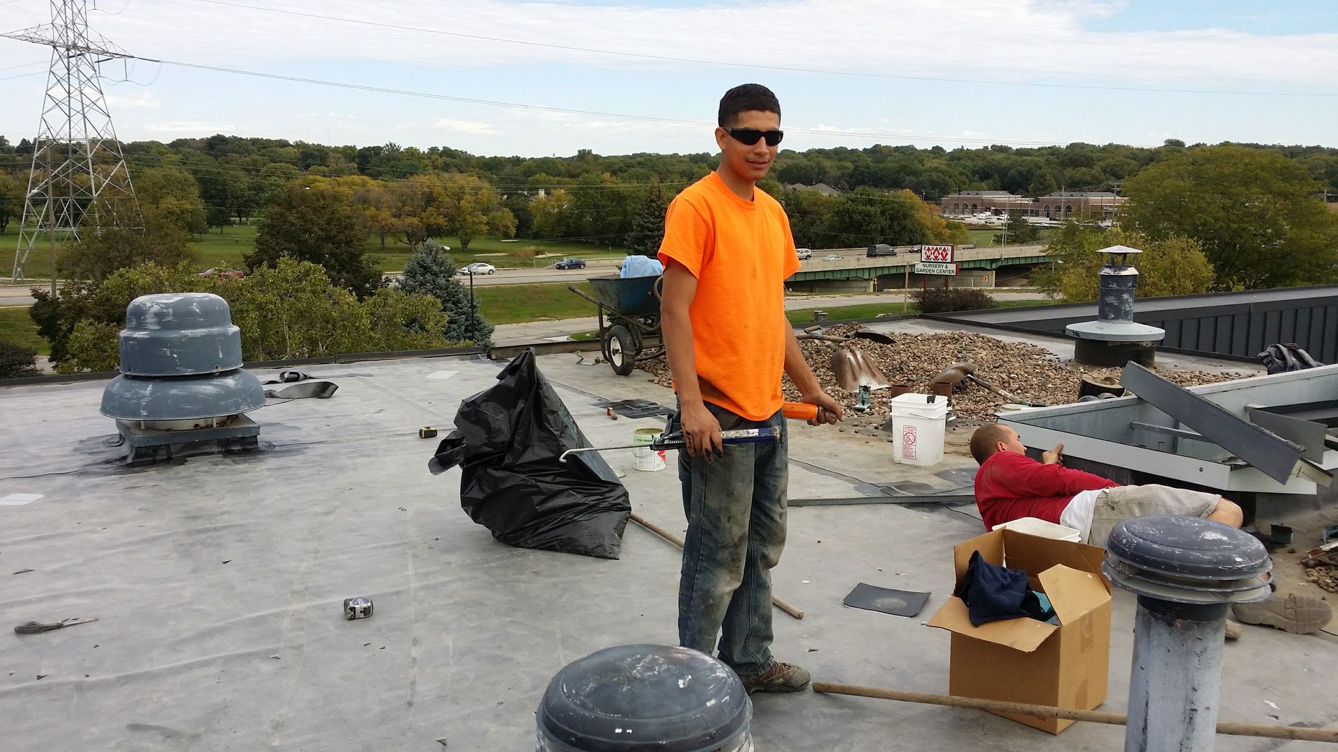 A worker in an orange shirt stands on a flat commercial roof with equipment, while another person works nearby.