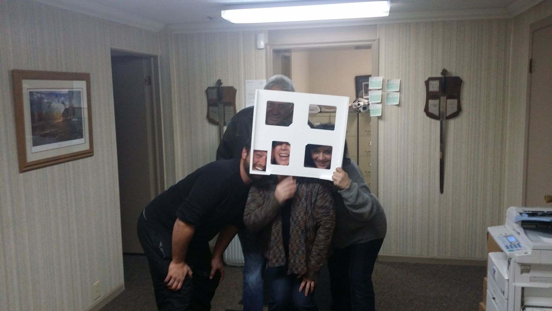 Three people in an office stand together behind a poster with four cutouts, showing their faces through the openings.