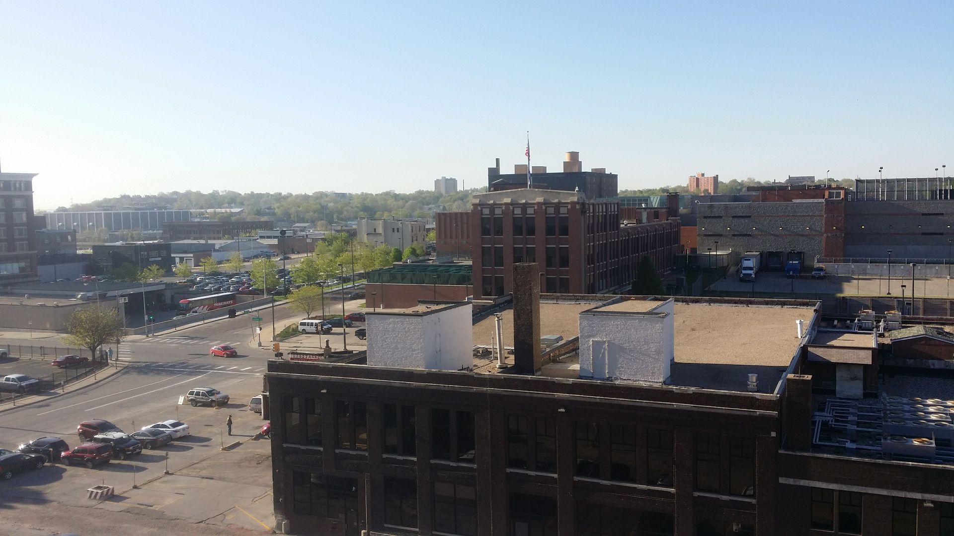 A high-angle view of a city landscape featuring old brick buildings, a parking lot, and distant hills under a clear sky.