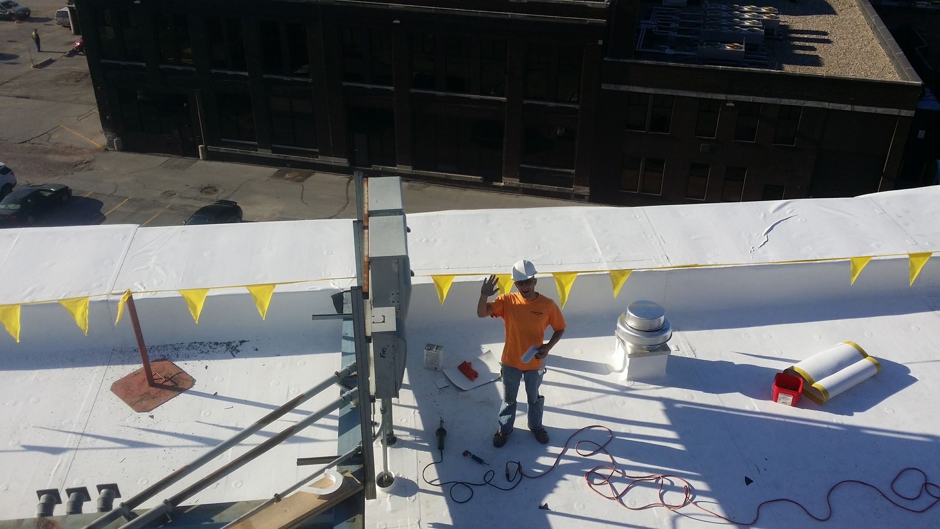 A worker in a bright orange shirt and white hard hat stands on a flat white roof, gesturing near industrial equipment.