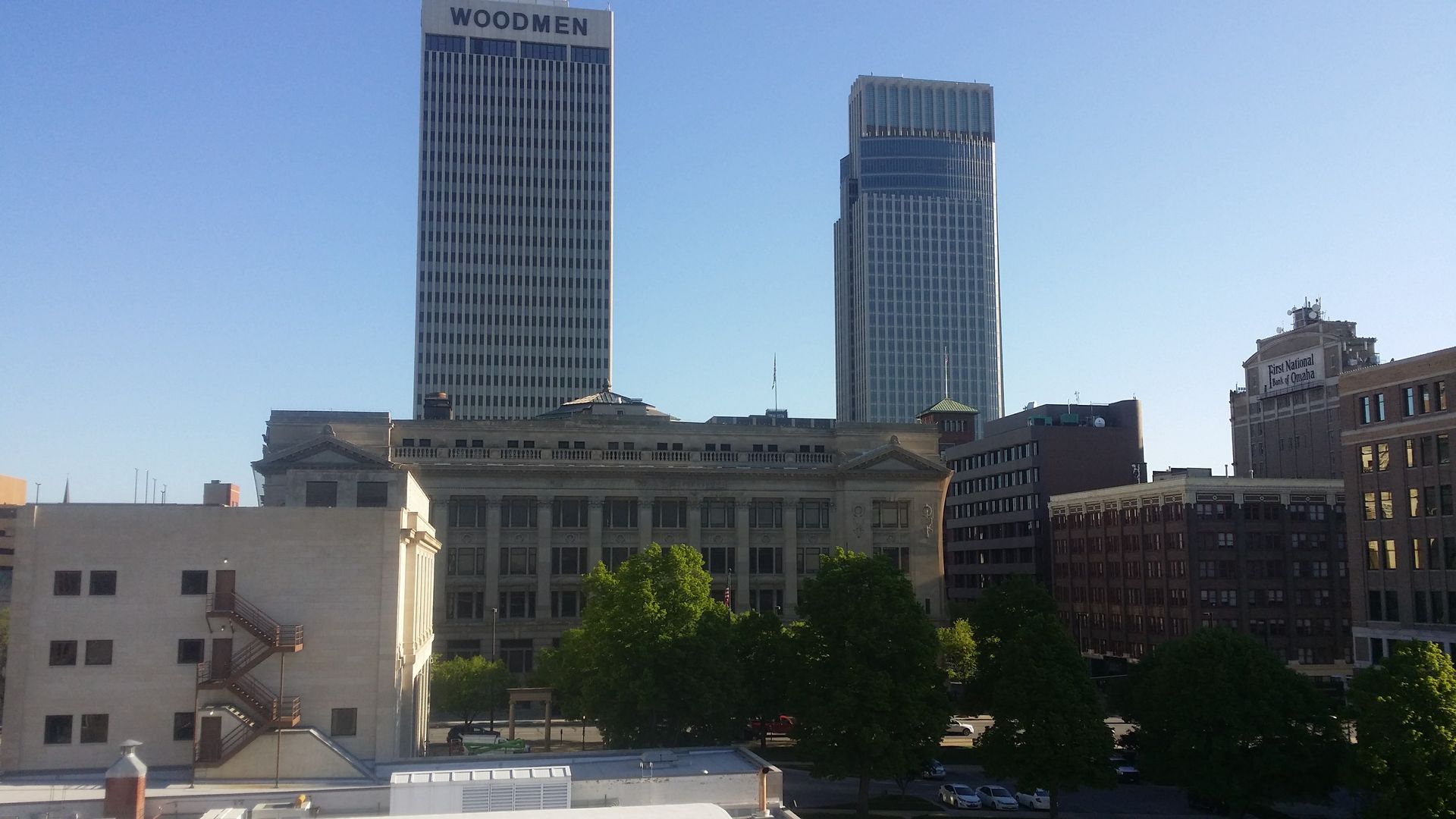 A cityscape featuring two prominent skyscrapers towering over older, lower-rise buildings and green trees under a clear sky.