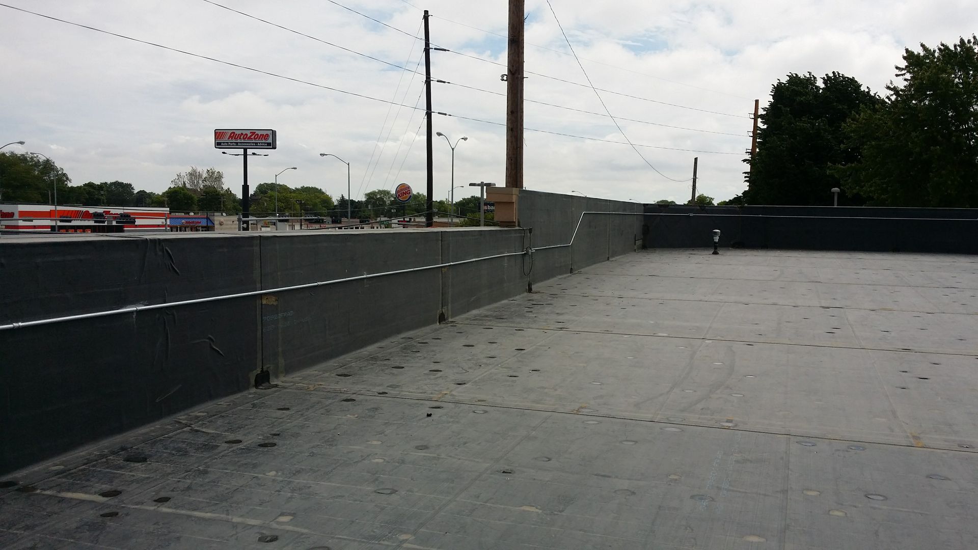 A flat, gray rooftop with a dark perimeter wall, showing construction markings and a distant commercial parking lot.