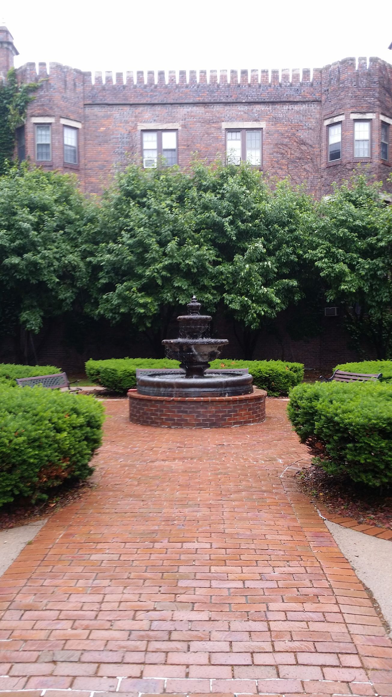 A brick walkway leads to a tiered stone fountain in a courtyard set against a historic stone building with lush hedges.