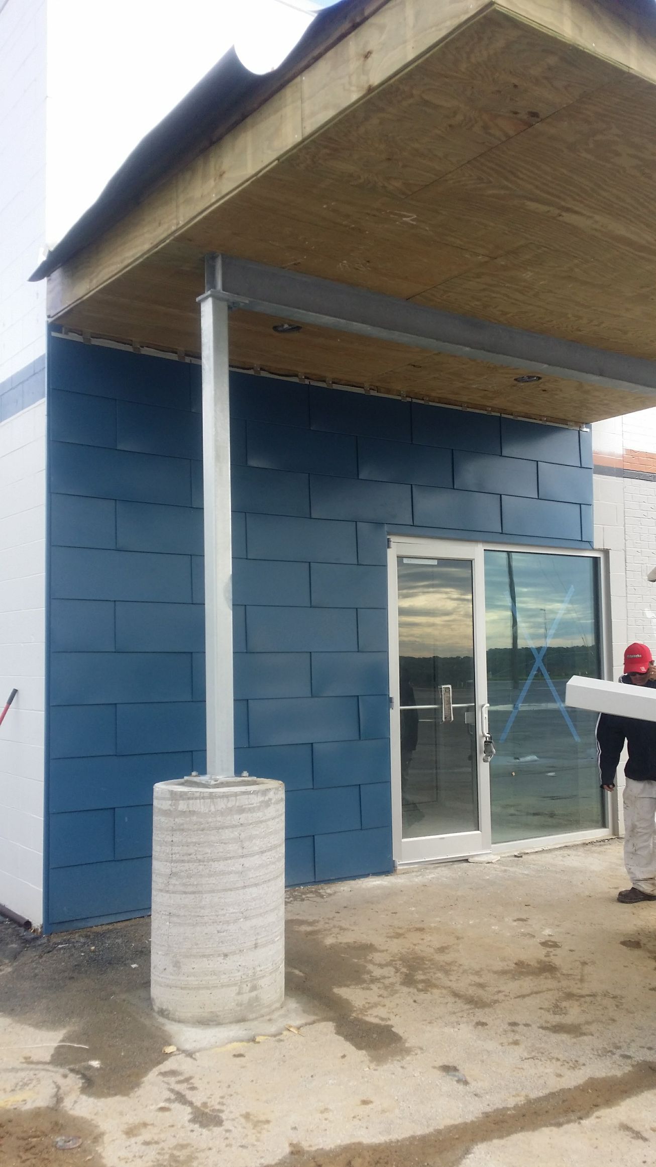 A person stands beside a commercial building entrance with dark blue block-patterned siding and a metal support pillar.