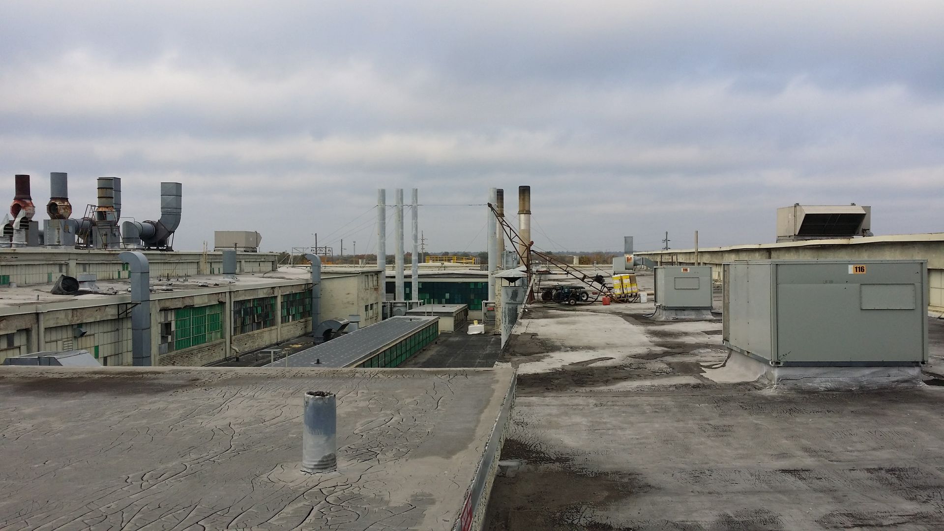 A wide view of a flat industrial rooftop under a cloudy sky, featuring HVAC units, pipes, and ventilation stacks.
