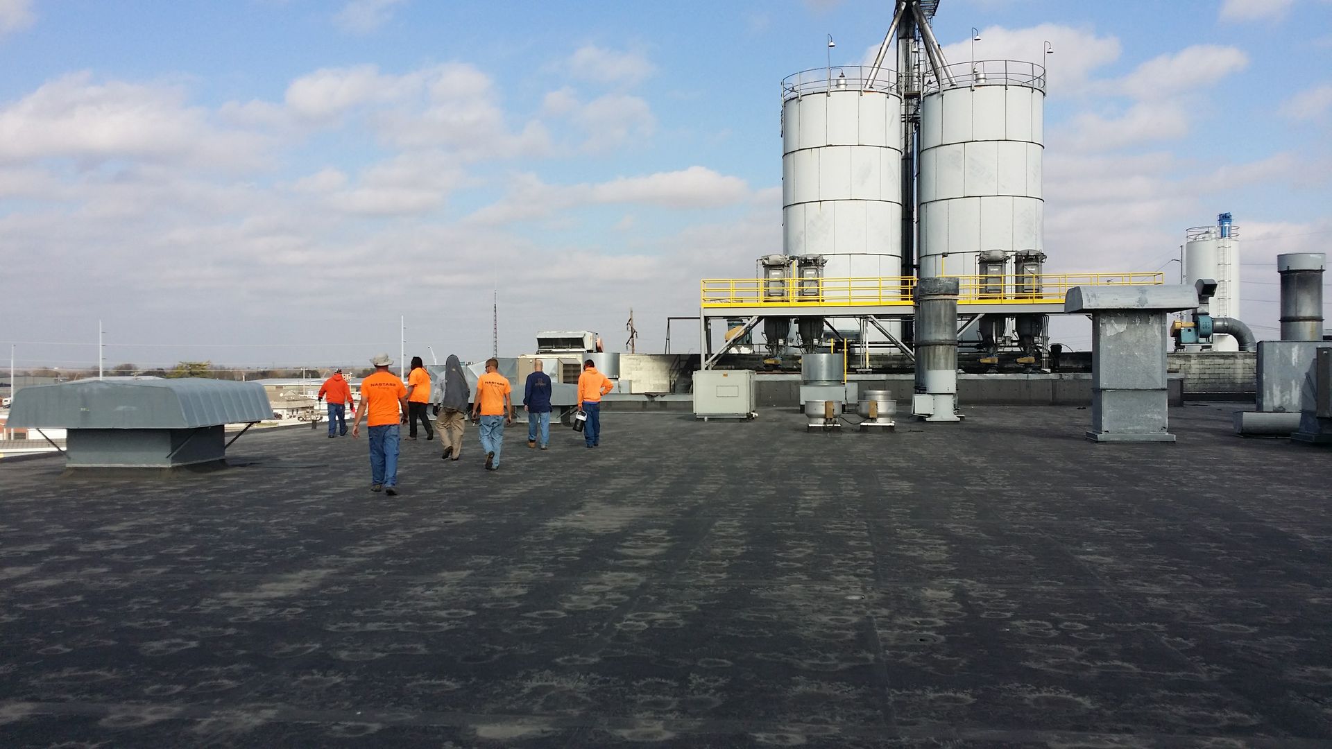 A group in high-visibility orange vests walking across a flat industrial rooftop toward large metal silos.