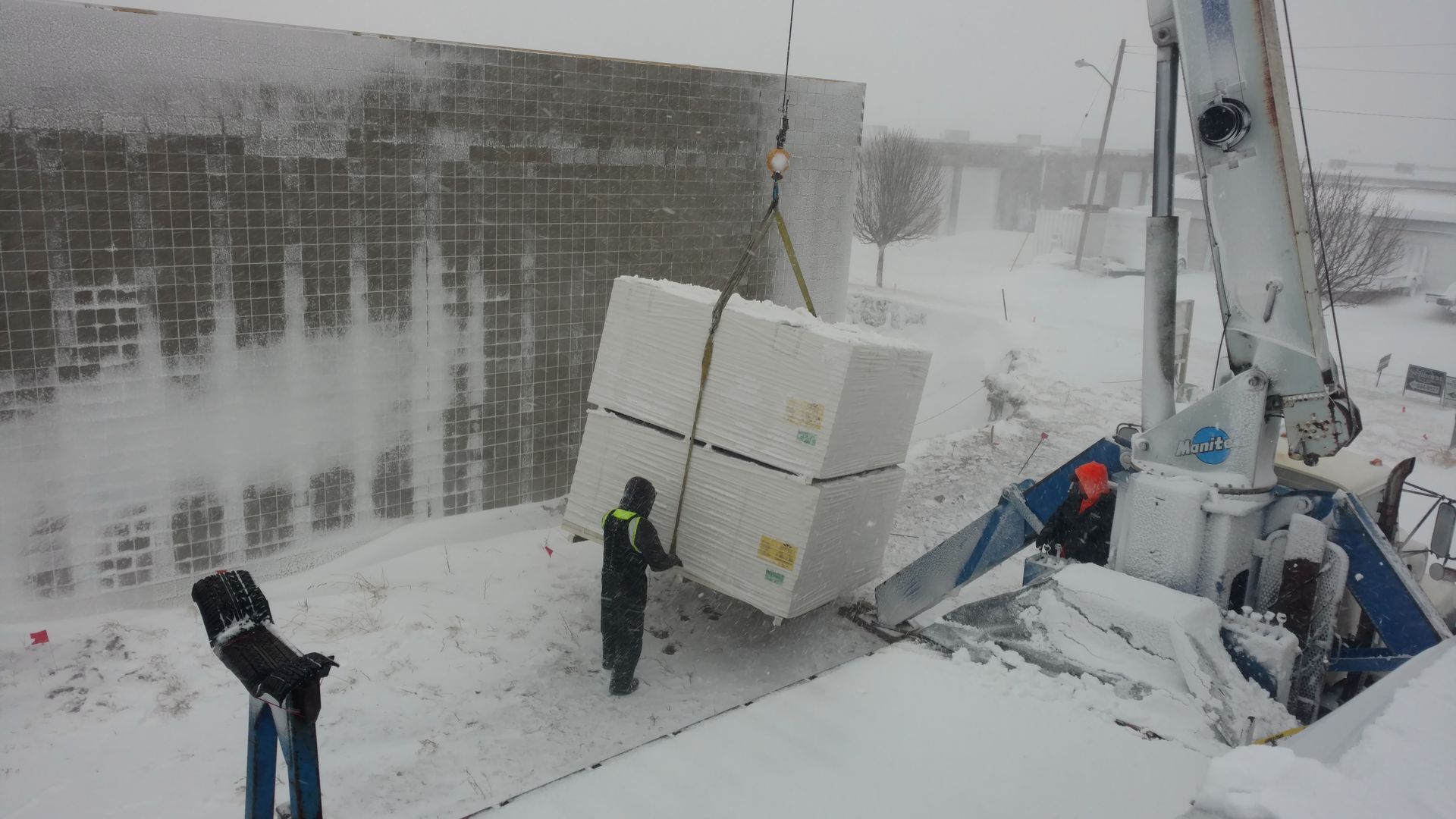 A crane lifts a large white cube in a snowy construction site, with workers nearby guiding the load.