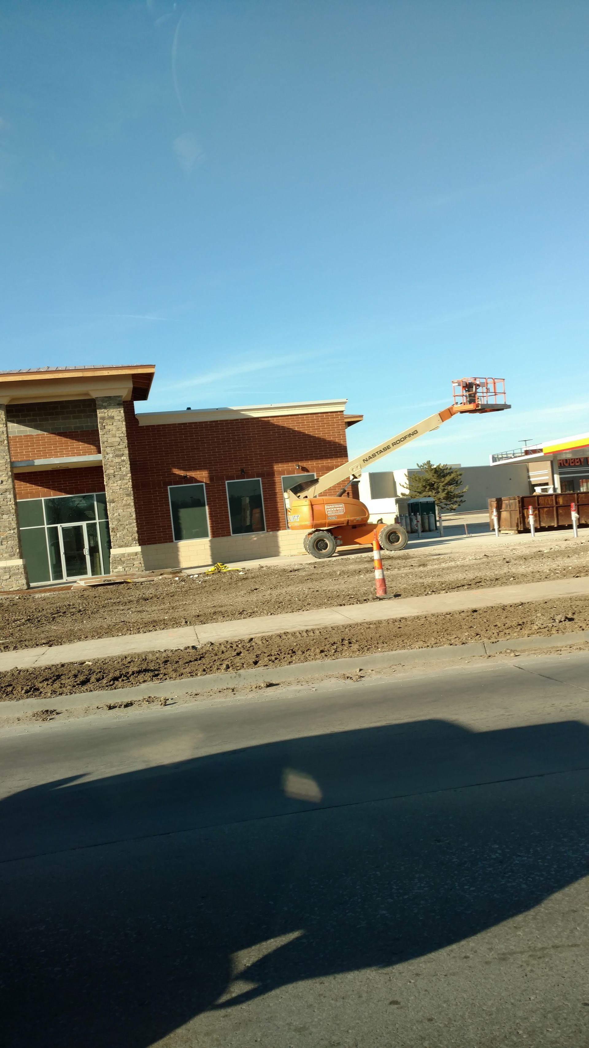 A construction boom lift extends its arm toward a partially built stone and siding building under a clear blue sky.