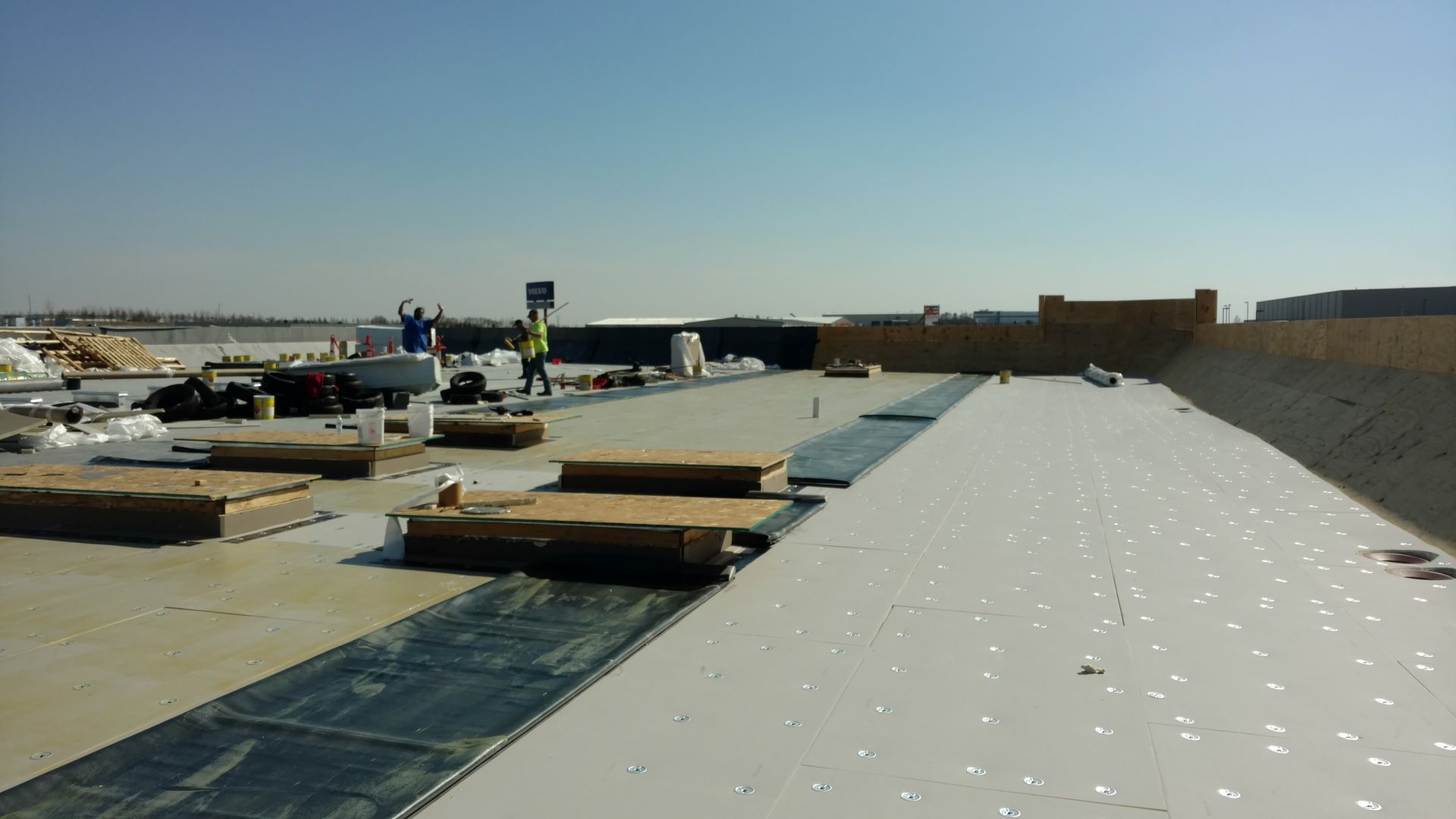 Workers installing white roofing material and insulation boards on a large, flat building rooftop under a clear blue sky.