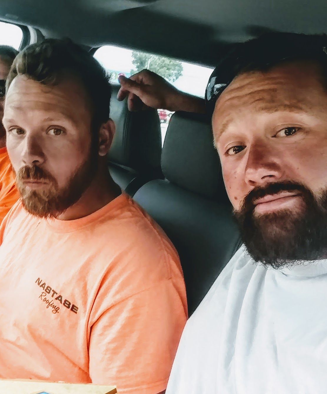Two people with beards, one in an orange shirt and one in a white shirt, sitting inside a vehicle.