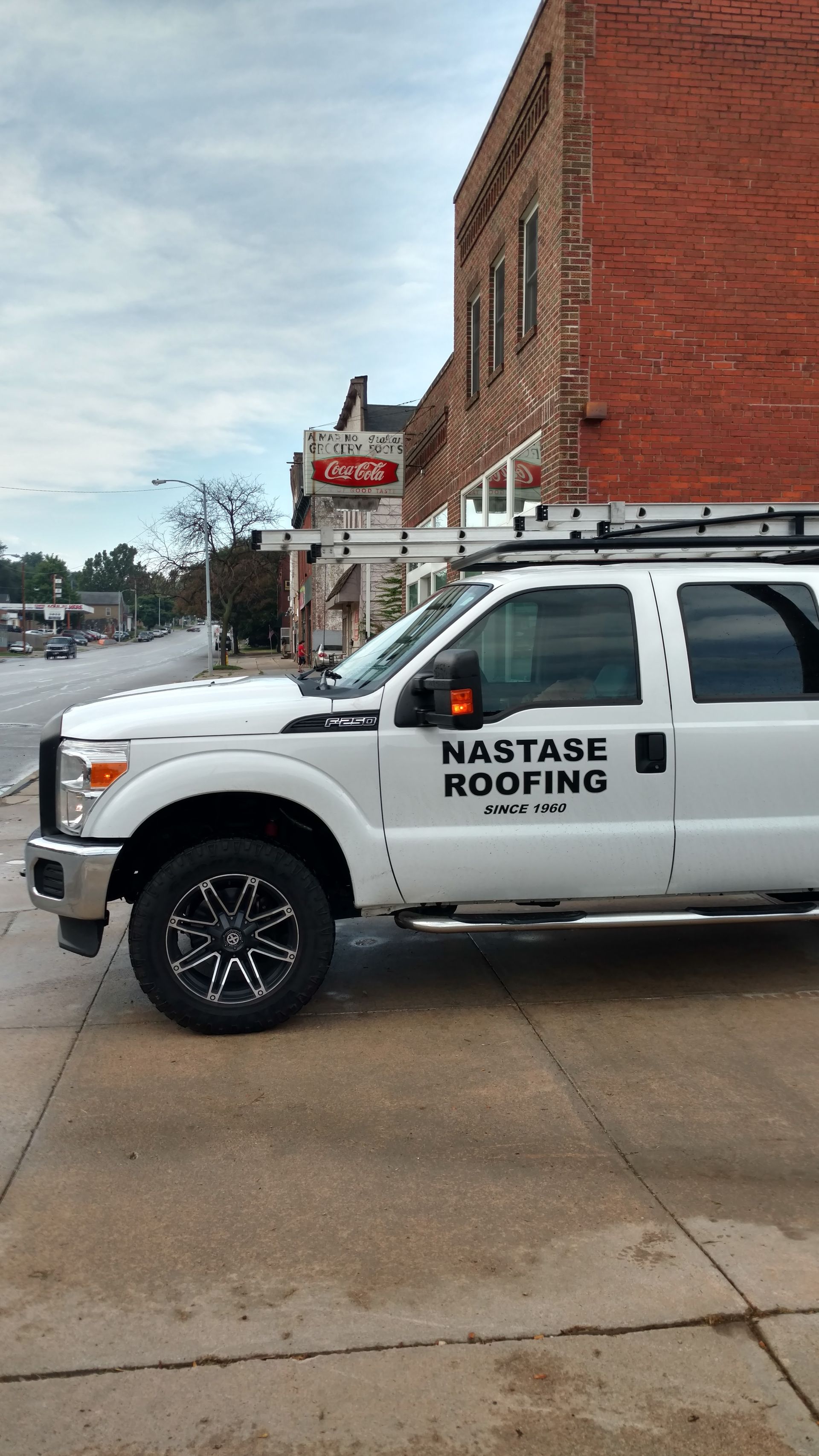 A white Nastase Roofing pickup truck parked on a street in front of a red brick building.