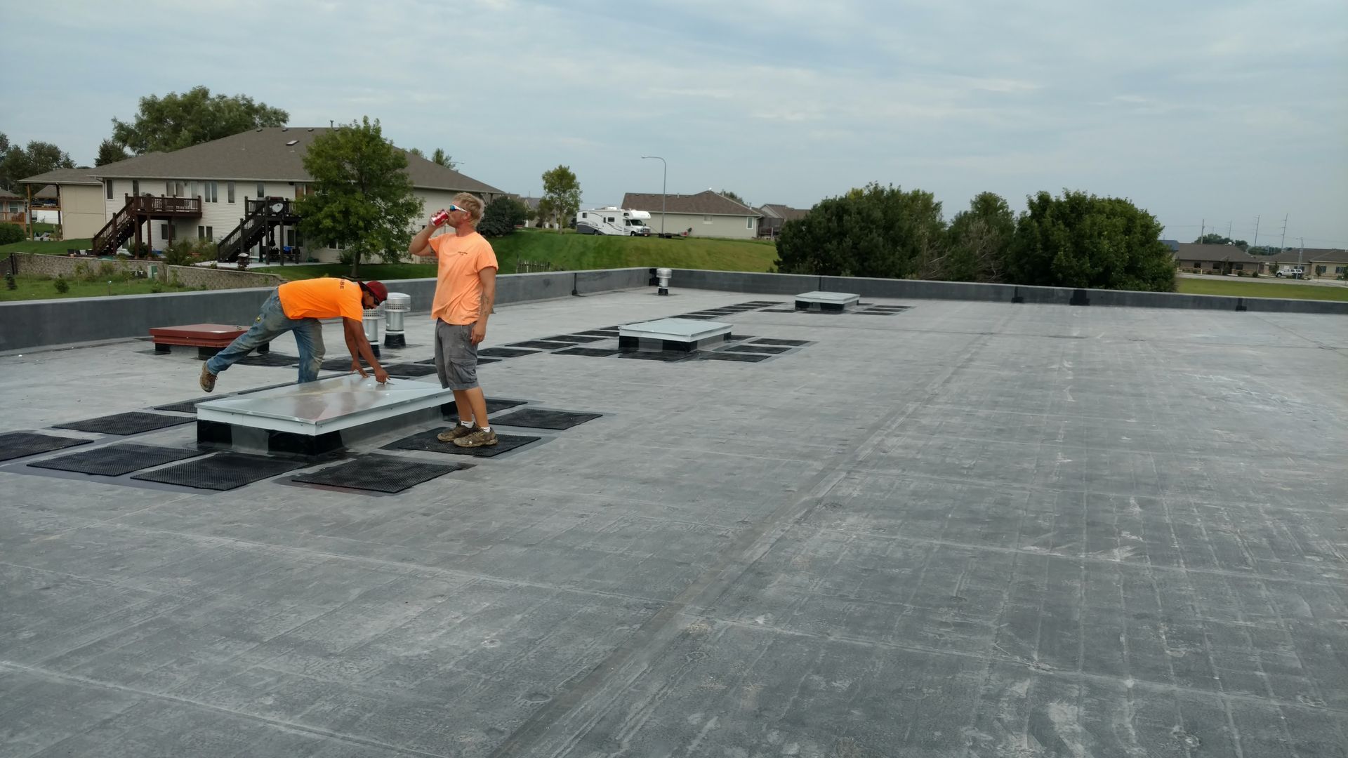 Two people in orange shirts work on a flat gray roof installing or repairing several skylights.