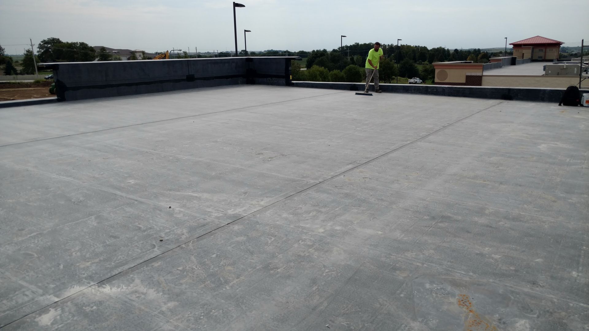 A construction worker in a high-visibility vest stands on a large, flat rooftop covered in grey, sheet-rolled material.